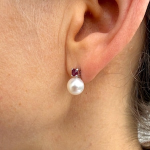 Peut inclure: Gros plan d'une boucle d'oreille à clou en perle avec une petite pierre précieuse rouge carrée sertie au-dessus de la perle. La boucle d'oreille est sertie dans un métal argenté. La boucle d'oreille est portée à l'oreille d'une personne.