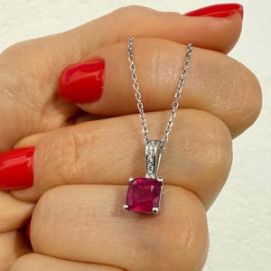 May include: A silver necklace with a square, deep red gemstone pendant. The pendant is set in a silver frame with small, clear accent stones. The chain is delicate and silver. The background is a light color.