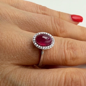 Può includere: Un anello d'argento con una grande pietra preziosa di rubino ovale, circondata da piccole pietre preziose trasparenti. Il rubino è di un colore rosso intenso ed è incastonato su una fascia sottile.