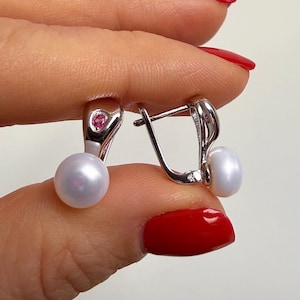 Puede incluir: Pendientes de plata con una perla blanca y una pequeña gema rosa. Los pendientes están sostenidos por una mano con esmalte de uñas rojo. Cierre de palanca.