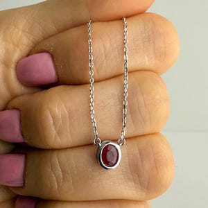 May include: A silver necklace with a small, oval ruby gemstone. The ruby is set in a bezel setting and suspended from a fine chain. The necklace is held in a hand with pink nail polish.