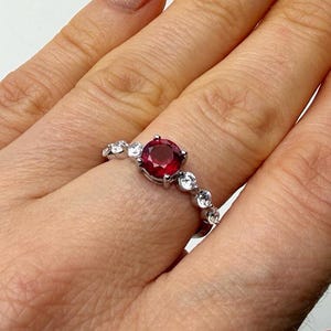 Può includere: Un anello d'argento con una gemma rosso scuro rotonda al centro, circondata da gemme chiare più piccole. L'anello è indossato su un dito, evidenziando il suo design e la sua brillantezza. È un gioiello.