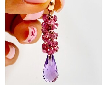 May include: A rose gold earring with a cluster of pink gemstones and a large, faceted, teardrop-shaped purple gemstone. The earring is held up by a hand with pink nail polish.