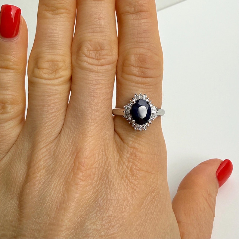 Natural blue sapphire engagement ring held between two fingers, alternating baguette and round white stones forming a structured halo