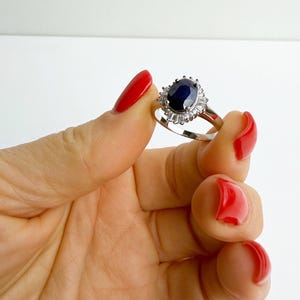 Hand holding a natural sapphire halo ring at eye level, the band reflecting light and the blue stone appearing nearly black at center