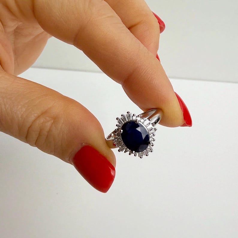 Oval sapphire engagement ring with radiating white stone halo worn on the middle finger, band visible curving around the finger