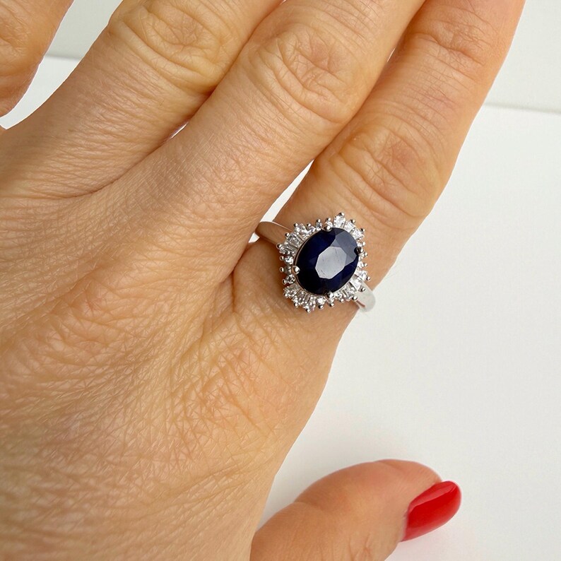 Hand holding a sterling silver ring with a deep blue oval sapphire in a ballerina halo, stones radiating outward in a crown pattern