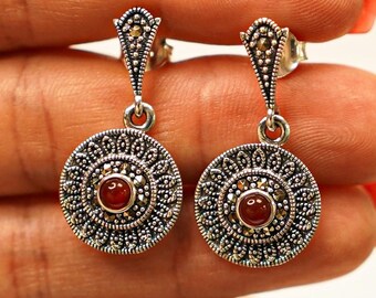 May include: A pair of silver drop earrings. Each earring has a circular pendant with a central red gemstone and detailed patterns. The earrings have a decorative top section with small, gold-coloured accents.
