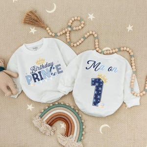 May include: Two white long-sleeved baby bodysuits. One features "Birthday PRINCE" in blue and gold, the other has "Ma on" and a number "1" with a crown and star pattern. A rainbow and wooden beads are also in the image.