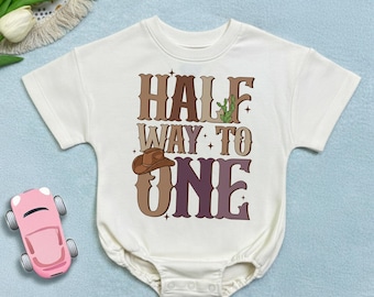 May include: Cream-colored baby bodysuit with the text "HALF WAY TO ONE" in brown and purple lettering. A cowboy hat and cactus graphic are included in the design. The bodysuit has short sleeves and a snap closure.