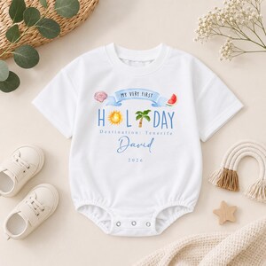 May include: A white short-sleeved baby bodysuit featuring a graphic print with the text "MY VERY FIRST HOLIDAY" on a blue banner, "Destination: Tenerife", the name "David", and the year "2026". The design includes a seashell, sun, palm tree, and watermelon.