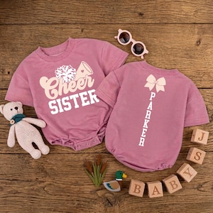 May include: Two mauve baby rompers. One says "Cheer Sister" with a cheerleader graphic. The other says "Parker" vertically with a bow graphic. Accessories include sunglasses, wooden blocks, and a teddy bear.