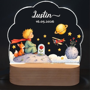 Peut inclure: Une veilleuse en acrylique personnalisée avec une illustration imprimée d'un jeune garçon aux cheveux blonds et d'un renard assis sur une lune, une rose rouge sous cloche, une planète jaune, un avion rouge et des étoiles. Texte : Justin 16.05.2026.