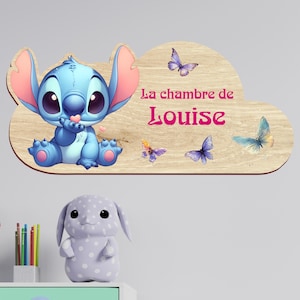 Peut inclure: Une plaque murale en bois en forme de nuage représentant un personnage bleu aux grandes oreilles avec des détails roses et quatre papillons colorés. Le texte "La chambre de Louise" est imprimé en rose sur la surface en bois clair.