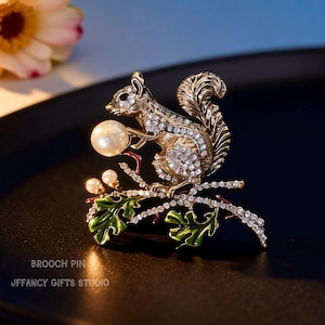 May include: A gold-toned squirrel brooch holding a pearl, perched on a branch with green leaves and sparkling accents. The brooch is embellished with clear crystals. Text on the image reads: BROOCH PIN, JEFFANCY GIFTS STUDIO.