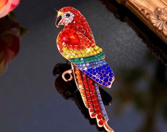 May include: A parrot-shaped brooch featuring a gold-tone metal frame encrusted with multi-colored rhinestones. The bird design displays a gradient of red, orange, yellow, green, and blue crystals, with a larger red stone on the wing section.