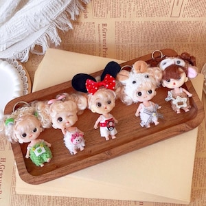 May include: A collection of miniature doll keychains with various outfits and accessories, including a Minnie Mouse-inspired design with a red bow. The dolls are arranged on a wooden tray, showcasing their detailed features and charm.