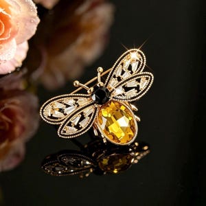 May include: A bee-shaped brooch featuring a large oval yellow crystal body, a round black crystal head, and wings outlined in gold metal with inlaid clear baguette-cut crystals. The piece has a reflective finish and a pin mechanism on the back.