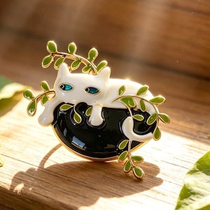 May include: A metal brooch featuring a white cat resting on a black, rounded base. The cat has bright blue eyes and is surrounded by gold-toned branches with small, vibrant green leaves that extend across the cat's body and the black base.