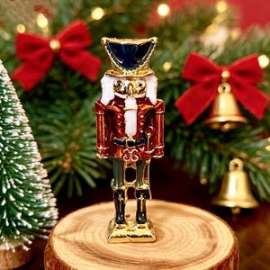 May include: A small, metallic nutcracker figurine featuring a glossy red uniform with gold accents, a dark blue hat, and white hair. The nutcracker stands upright on a rectangular base, showcasing intricate enamel detailing and a polished finish.