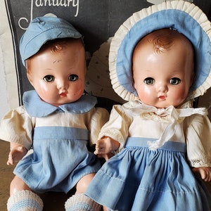 May include: Two vintage porcelain dolls dressed in blue and white outfits. The dolls have painted features and are wearing knitted socks.
