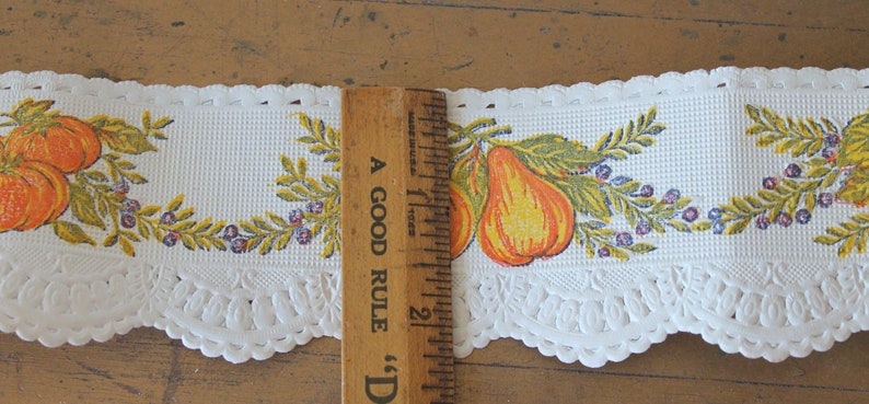 Puede incluir: Borde de encaje blanco con un borde festoneado y un dise&ntilde;o impreso de peras amarillas y naranjas con hojas verdes y bayas moradas. El borde se mide con una regla que dice "A Good Rule" y tiene 5 cm de largo.