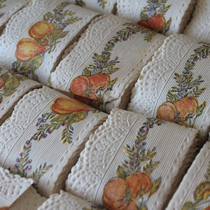 May include: A close-up of several rolls of white lace trim with a repeating pattern of green leaves, red berries, and orange fruit. The trim is perfect for adding a touch of vintage charm to any project.