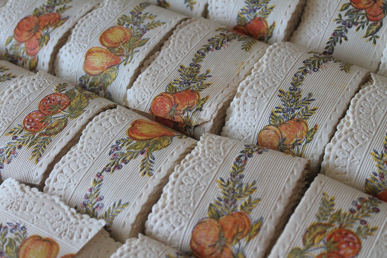 Puede incluir: Primer plano de varios rollos de encaje blanco con un patr&oacute;n repetido de hojas verdes, bayas rojas y frutas naranjas. El encaje es perfecto para agregar un toque de encanto vintage a cualquier proyecto.