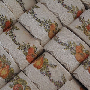 Puede incluir: Primer plano de varios rollos de encaje blanco con un patr&oacute;n repetido de fruta naranja y amarilla, hojas verdes y peque&ntilde;as bayas moradas.