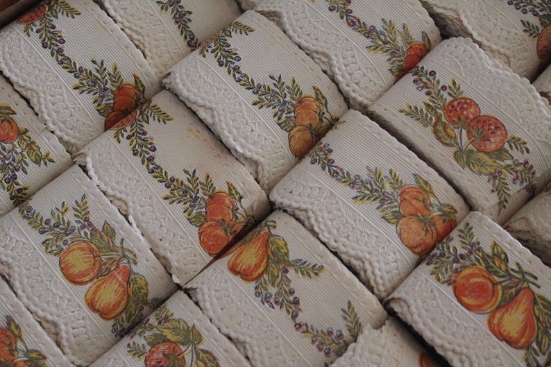 Puede incluir: Primer plano de varios rollos de encaje blanco con un patr&oacute;n repetido de fruta naranja y amarilla, hojas verdes y peque&ntilde;as bayas moradas.