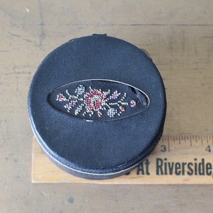 May include: A black round box with a floral design embroidered on the top. The design features a red rose with white and silver accents.