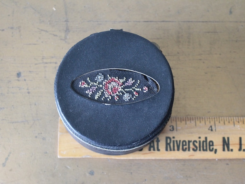 May include: A black round box with a floral design embroidered on the top. The design features a red rose with white and silver accents.
