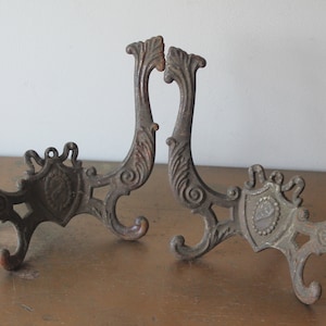 May include: Two ornate metal hooks with a decorative shield design. The hooks are made of cast iron and have a rustic brown finish.