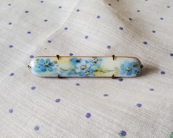 Puede incluir: Un broche de porcelana vintage con un delicado diseño de flores azules de nomeolvides sobre un fondo blanco.