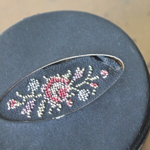 May include: A black round box with a fabric oval cutout featuring a colourful floral design embroidered with small beads.