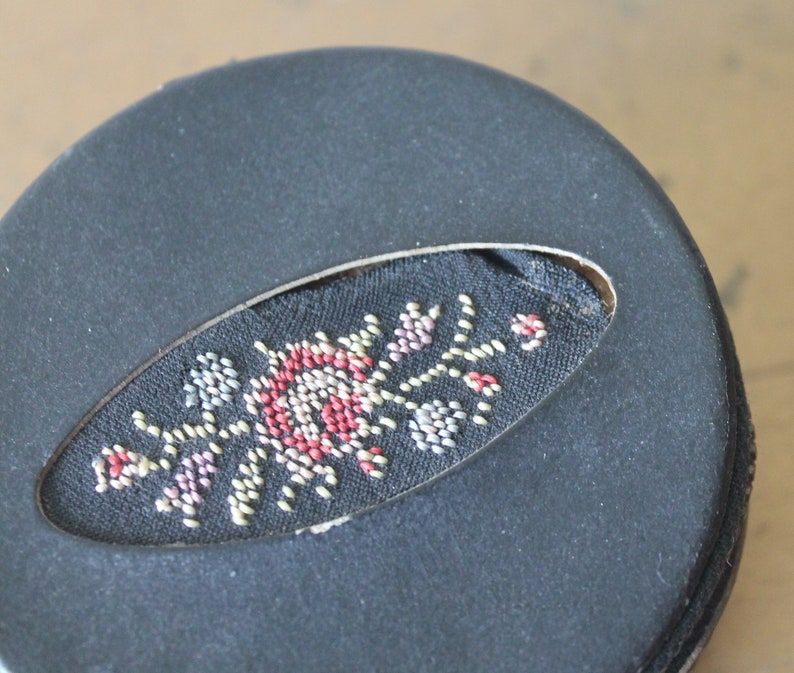 May include: A black round box with a fabric oval cutout featuring a colourful floral design embroidered with small beads.