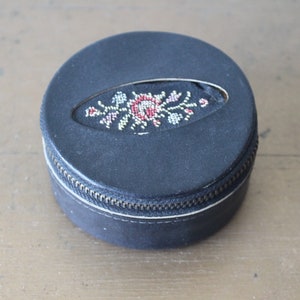 May include: A small, round, black leather trinket box with a zip closure. The lid features a floral design embroidered in red, white and gold thread.