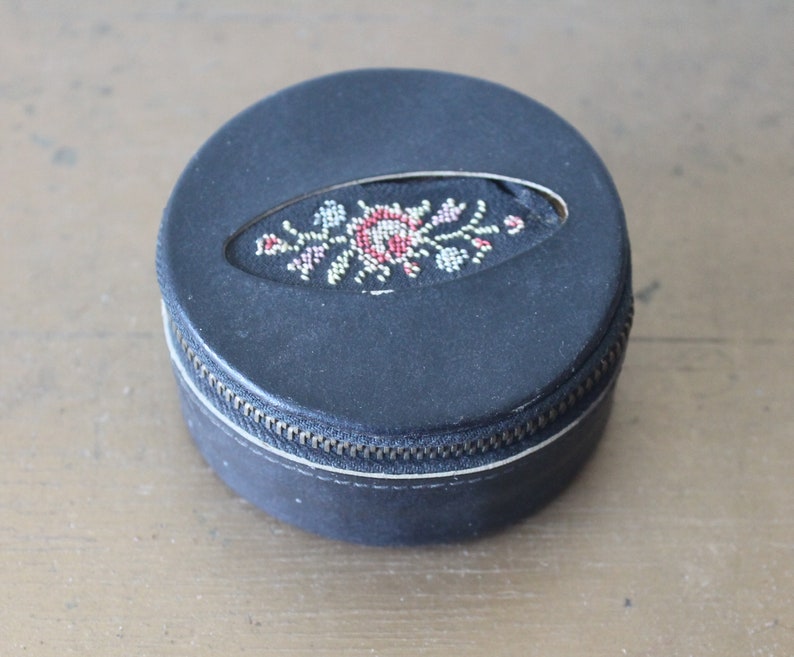 May include: A small, round, black leather trinket box with a zip closure. The lid features a floral design embroidered in red, white and gold thread.