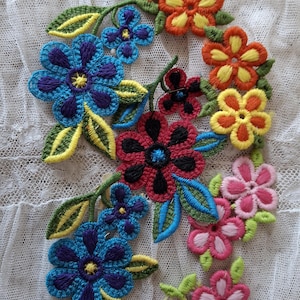 May include: Embroidered floral applique in a variety of colors, including blue, red, orange, and pink. The flowers are detailed with yellow and black accents, and green leaves. The applique is set against a white lace background.