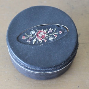 May include: A round, black leather trinket box with a zip closure. The lid features a floral design embroidered in red, white and gold thread.