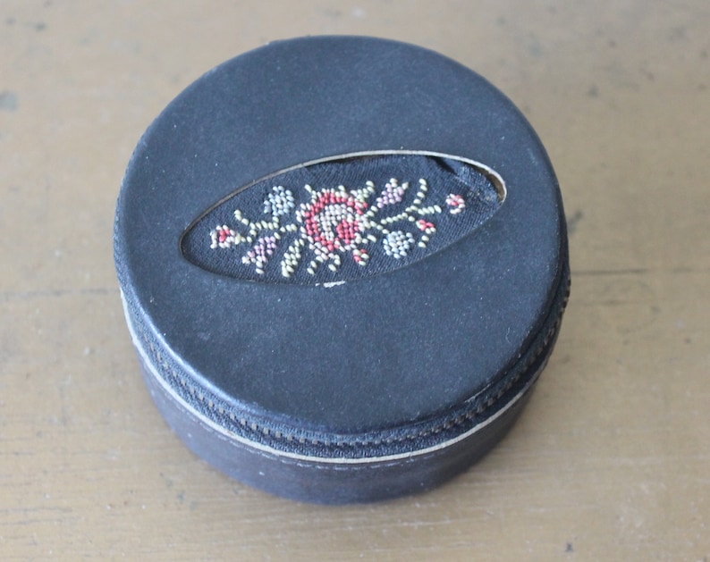 May include: A round, black leather trinket box with a zip closure. The lid features a floral design embroidered in red, white and gold thread.