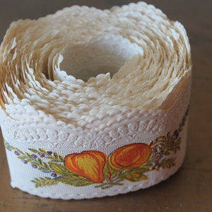 Puede incluir: Un rollo de encaje blanco con un borde festoneado y un dise&ntilde;o floral con frutas naranja y amarillas. El encaje est&aacute; enrollado y listo para ser usado para manualidades o decoraci&oacute;n.