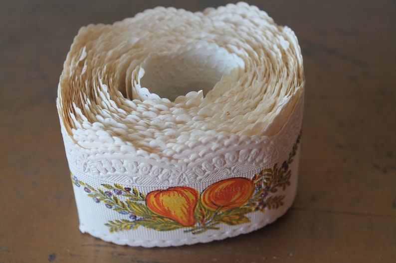 Puede incluir: Un rollo de encaje blanco con un borde festoneado y un dise&ntilde;o floral con frutas naranja y amarillas. El encaje est&aacute; enrollado y listo para ser usado para manualidades o decoraci&oacute;n.
