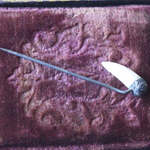 May include: A white bone pin with a silver filigree design on the end. The pin is on a dark purple velvet background.