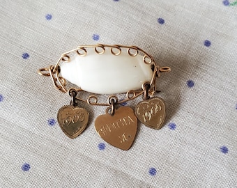 Puede incluir: Un broche dorado con una concha blanca y tres dijes en forma de corazón. Los dijes están grabados con las fechas "1905" y "1905" y la palabra "Virginia".