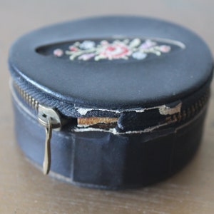 May include: A round black leather box with a zipper closure and a floral embroidery design. The box has a worn look with some of the leather peeling off.