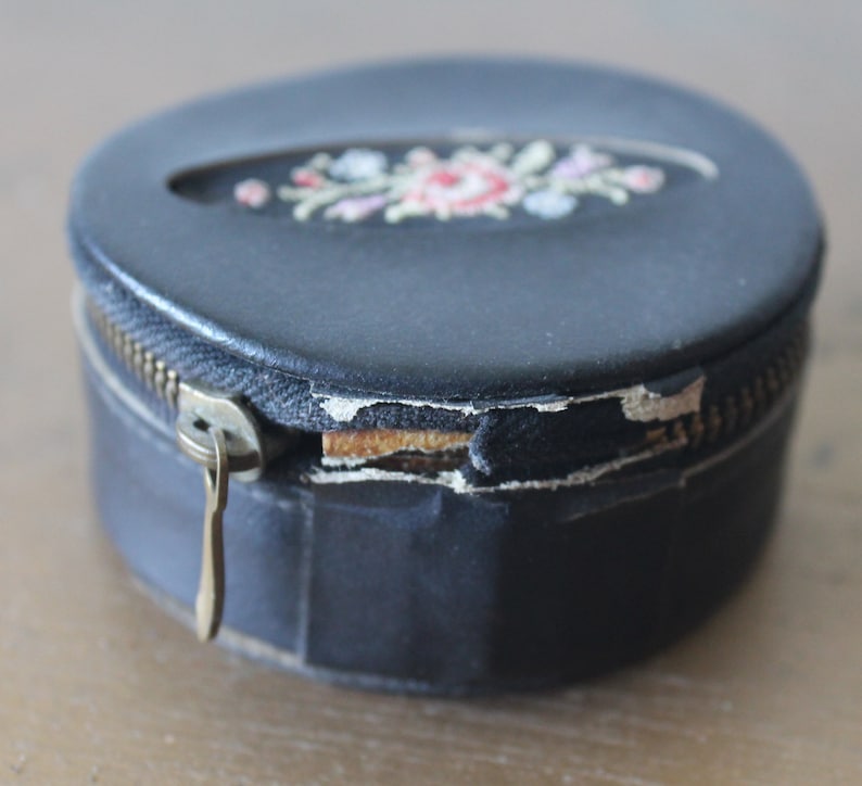 May include: A round black leather box with a zipper closure and a floral embroidery design. The box has a worn look with some of the leather peeling off.