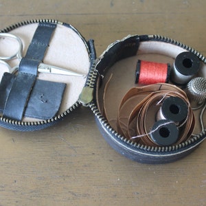 May include: A black leather sewing kit with a zip closure. The kit is open and contains a pair of scissors, a reel of red thread, a reel of white thread, a thimble, and a roll of brown thread.