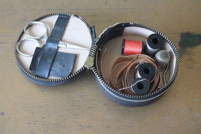 May include: A black leather sewing kit with a zip closure. The kit is open and contains a pair of scissors, a reel of red thread, a reel of white thread, a thimble, and a roll of brown thread.