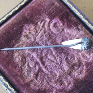 May include: A silver and white pin with a decorative floral design on the top. The pin is sitting in a velvet-lined box.